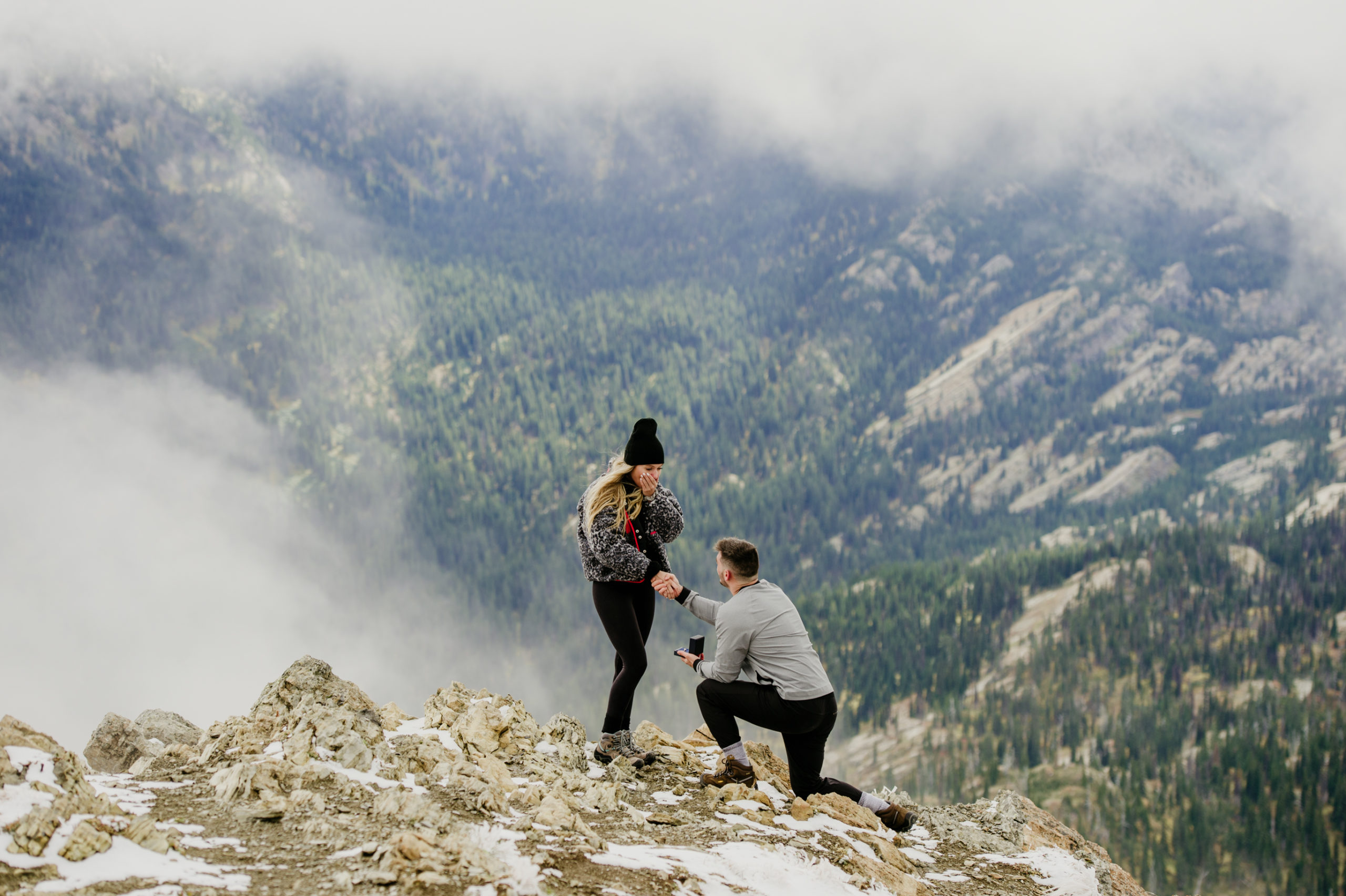 man proposing to his girlfriend on the top of the mountain while the clouds part