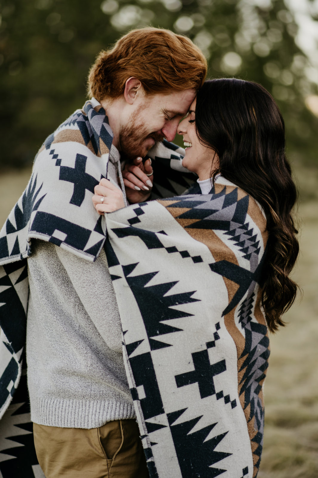 A red-haired man and raved haired woman's touch forwards, wrapped in a blanket, during their engagement photos in Missoula.