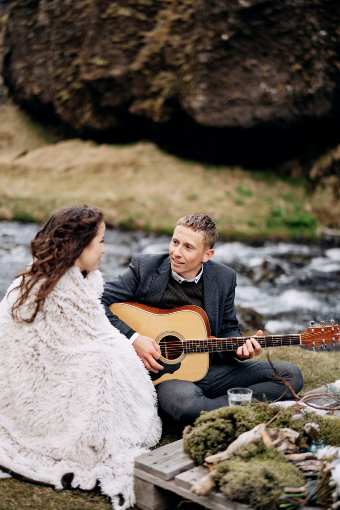 The wedding couple sits on the bank of a mountain river, at a table for a wedding dinner. The groom plays and sings for the bride.