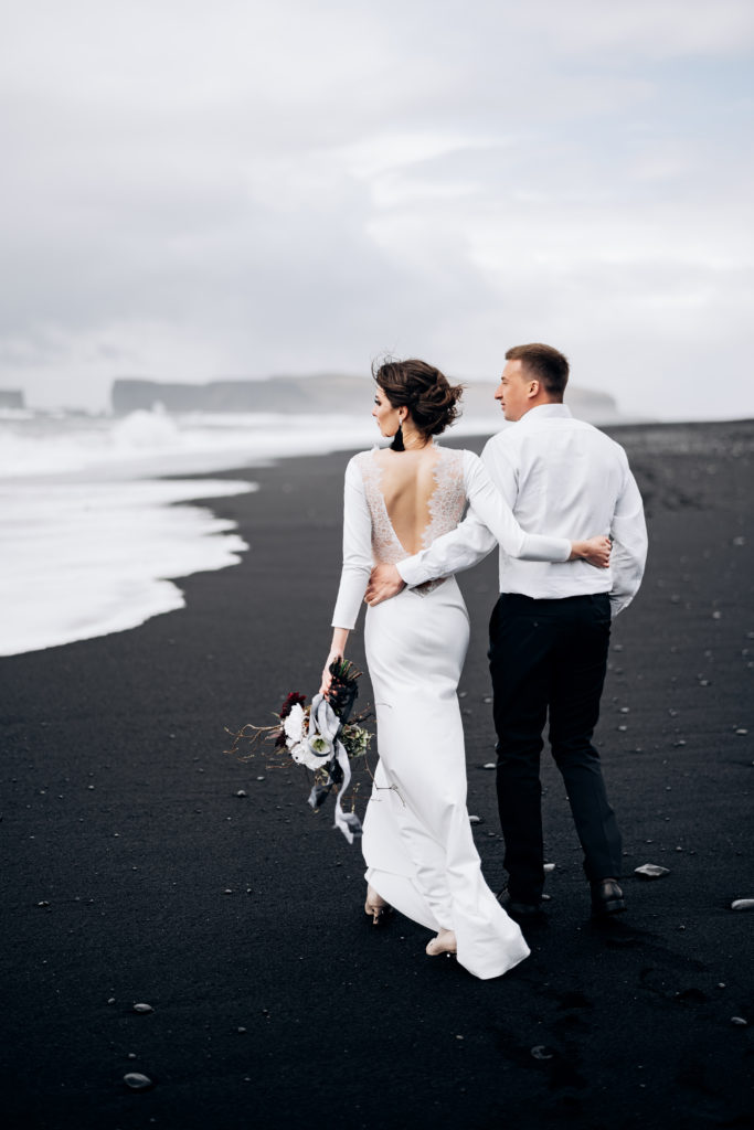 How to elope in Southern Iceland, top elopement location trends of 2022, Iceland Elopement, Wedding couple walks on black Beach Vic. A sandy beach with black sand on the shores of the Atlantic Ocean. Huge frothy waves. The groom hugs the bride.