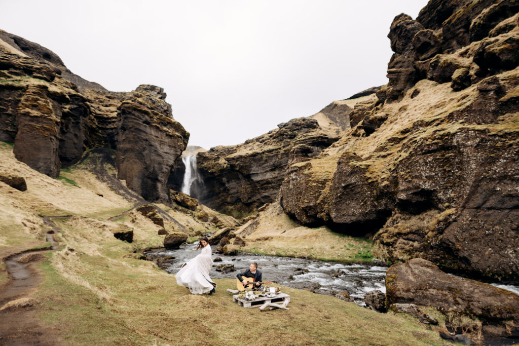 how to elope in Southern Iceland, top elopement location trends of 2022, Iceland Elopement. The groom plays the guitar sitting on the grass, at the improvised wedding table, the bride in a white dress and woollen plaid - dancing next to each other.