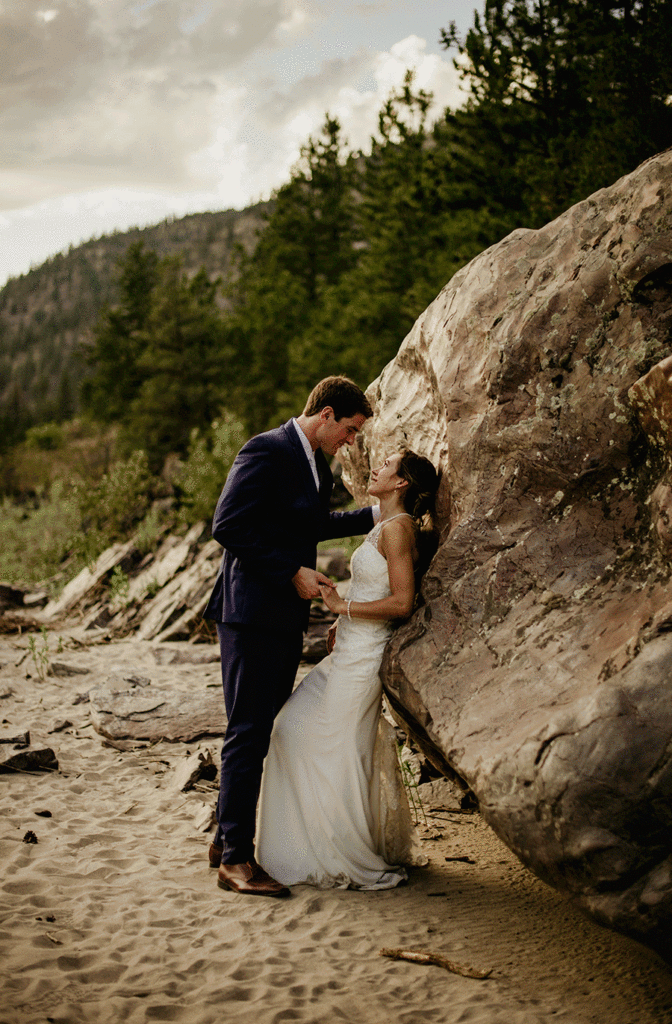 Our favorite wedding photography photos includes a bride with her back leaned up against a rock and a towering groom facing her and holding her hand. Trees in the distance.