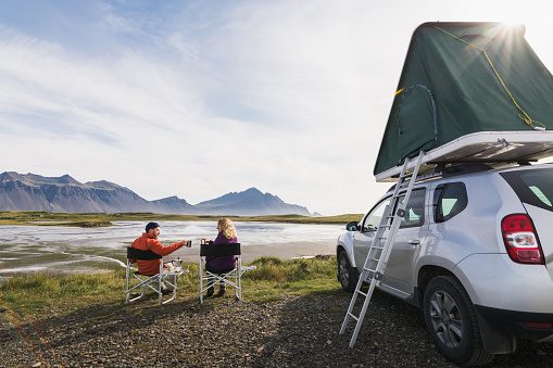 What are the benefits of eloping? We asked what are the reasons to elope from 20 of our real couples! Hofn, Iceland - August 2018: Young couple sitting in folding chairs next to offroad car with tent on the roof.