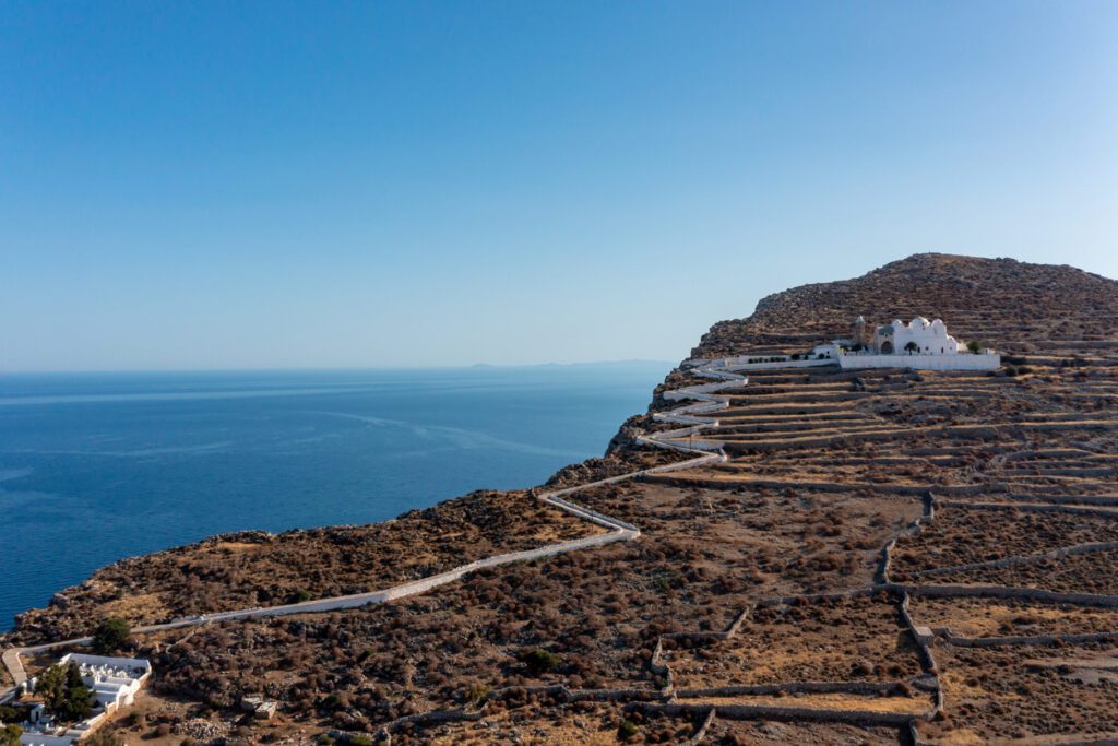 Folegandros island, Greece, Cyclades. Panagia Virgin Mary Church and long zigzag road aerial drone view. Traditional cycladic whitewashed church on top of a steep hill overlooking Chora and Aegean Sea