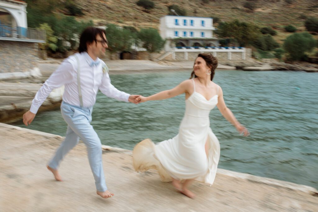 Couple running in Greece after packing for an adventure elopement. Bride in white dress, man in powder blue trousers and blue bow. Both barefoot.