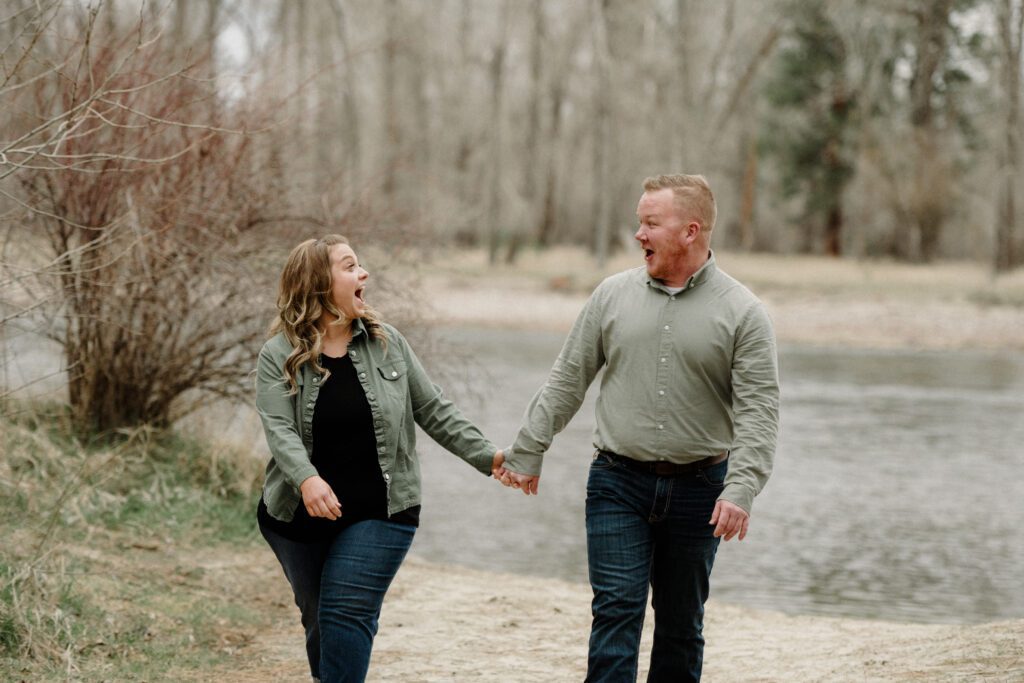 A couple laughs while looking at each other during engagement photos in Missoula. A river is behind them and trees are featured.