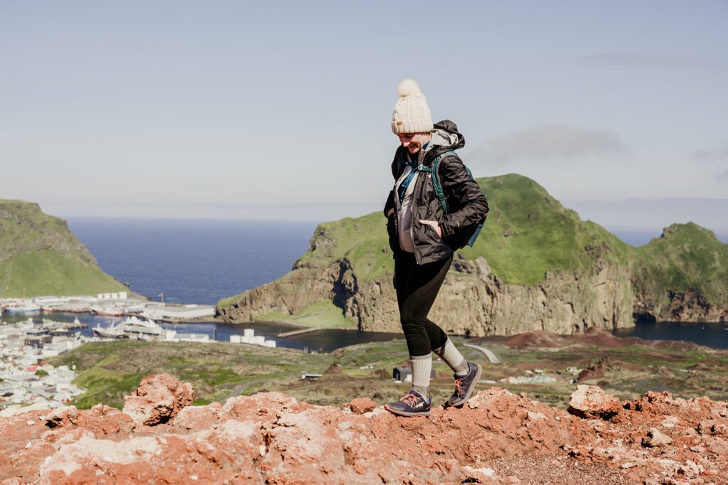 Photographer deep in thought about building up her associate team. Hiking in Iceland on red rocks with green hills and the ocean behind her. Dressed in winter hiking clothes.