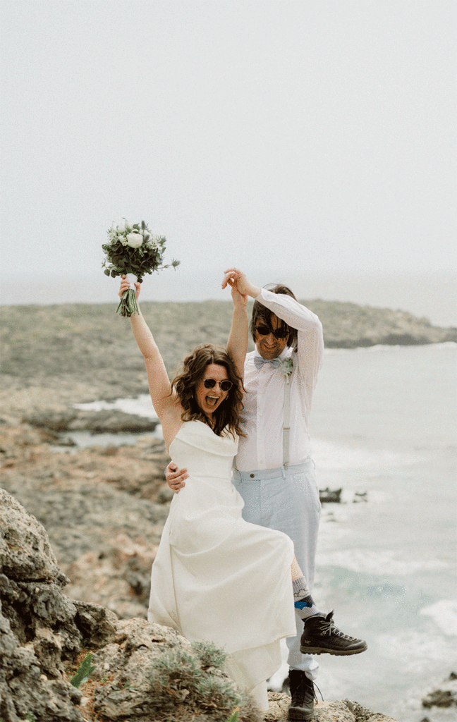 Scripts to tell Friends and Family that You’re Eloping! Photo of couple in Greece. Bride with her hands in the air, one with a bouquet, in excitement.