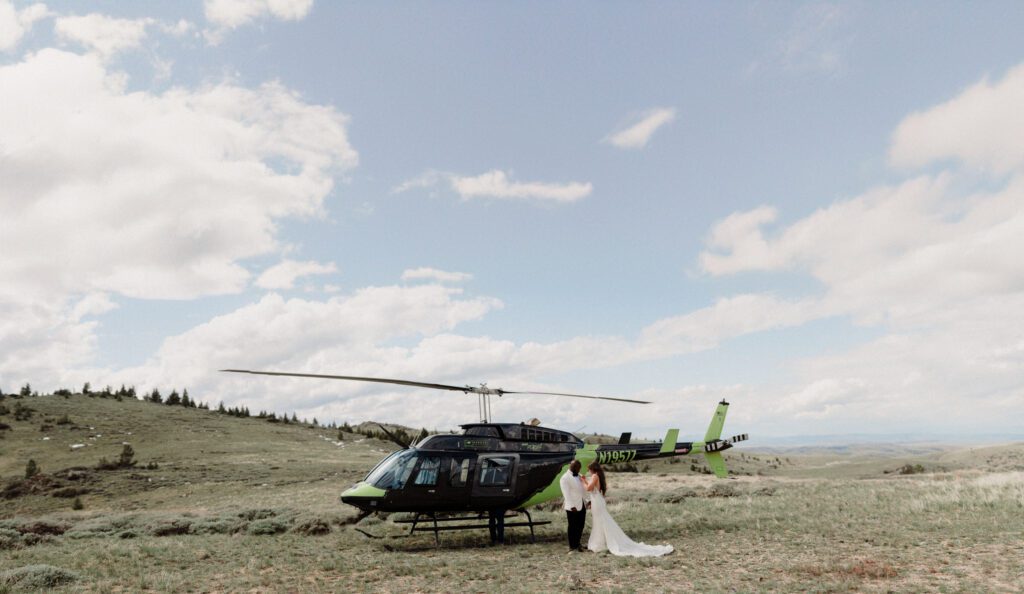 Guide to Booking 2 day elopements include having activities. Photo of bride and groom in wedding attire standing in front of a black and green helicopter.