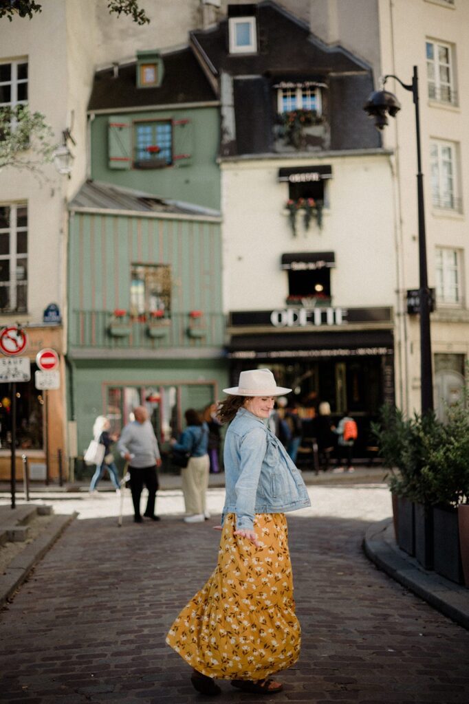 How to keep your sanity as a business owner. Business coach in the middle of busy European city. Wearing golden yellow floral dress, jean jacket, and white brimmed hat. Unbothered queen.