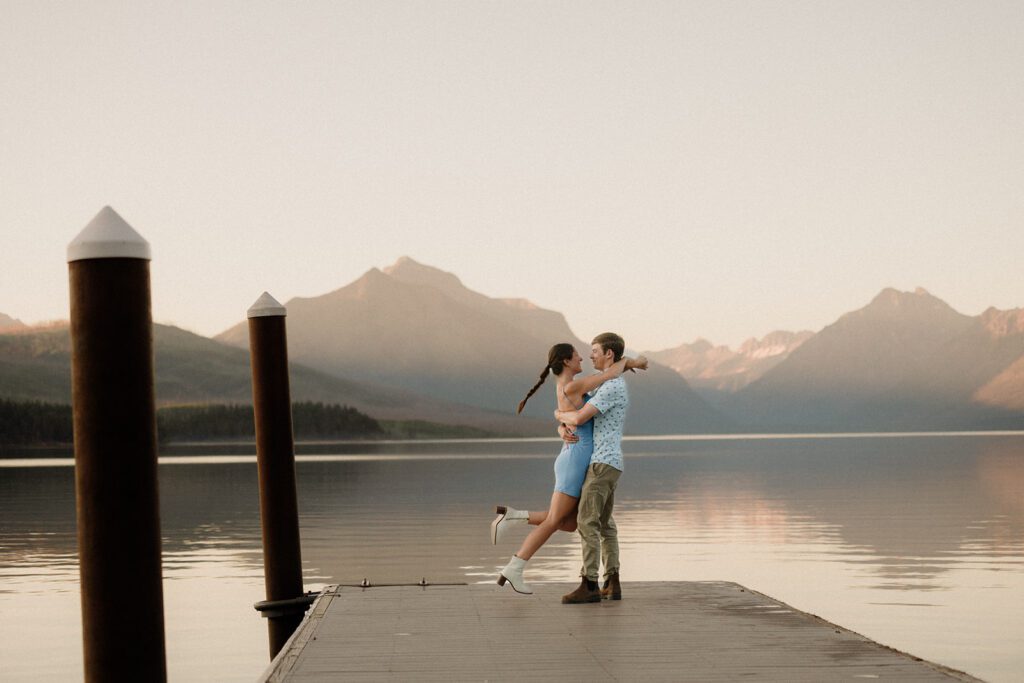 Woman jumping into man's arms on a dock with pastel mountains behind them.