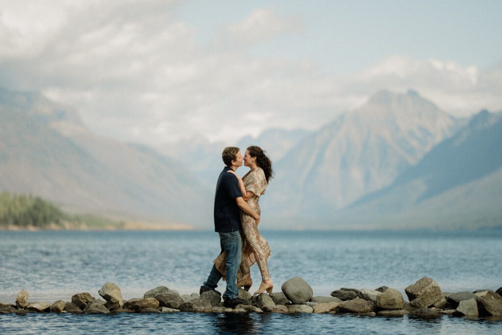 Top elopement destination: Glacier National Park. Couple kissing on rocks with water on either side of them. Mountains and clouds in the background