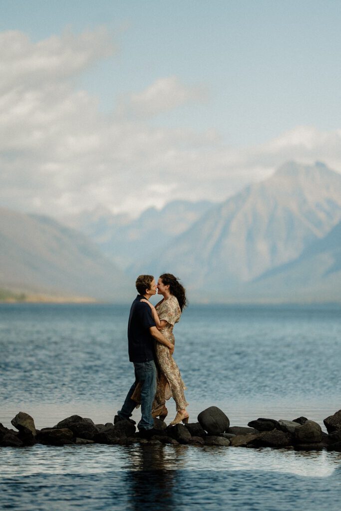 Adventure vox renewal at Glacier National Park with couple embracing on rocks, water on either side of them. 