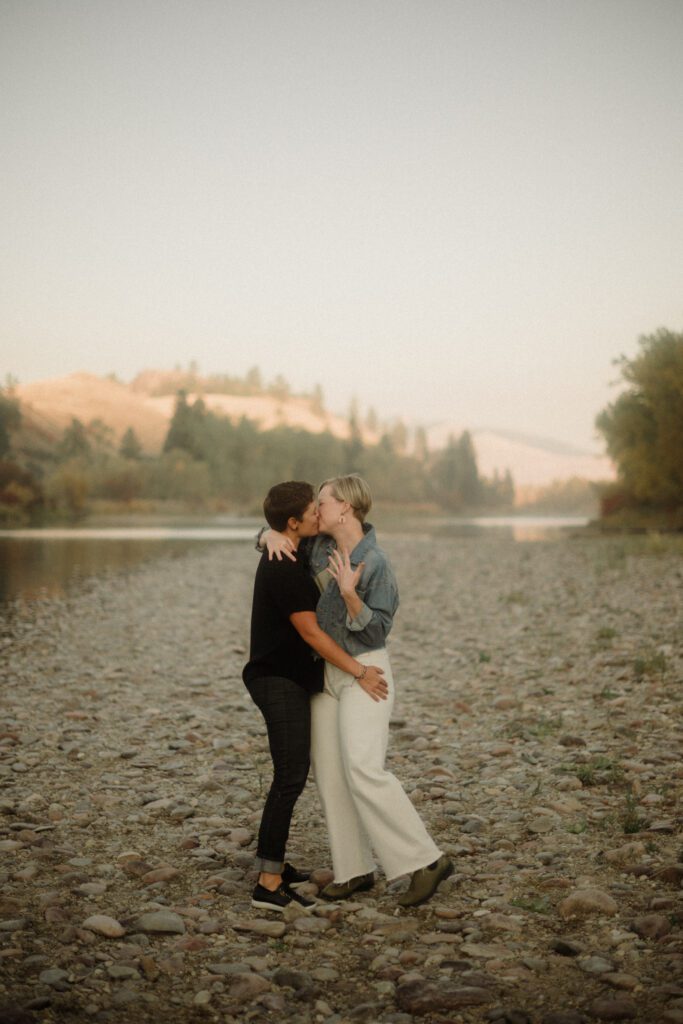 Lesbian Proposal in Missoula: Maclay Flat in Autumn. Photo of two women kissing after getting engaged. On the rocky shore line with trees surrounding them. 