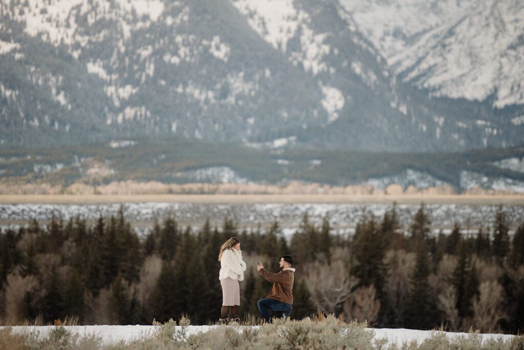 Proposal in Grand Teton. Man in brown  jacket on one knee with ring open to girlfriend. Grand Teton mountains behind them with trees.