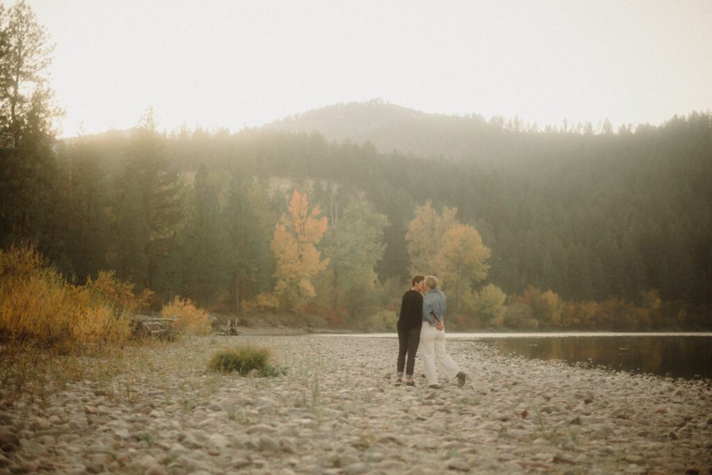 Lesbian Proposal in Missoula: Maclay Flat in Autumn. Photo of two women kissing after getting engaged. On the rocky shore line with trees surrounding them. 