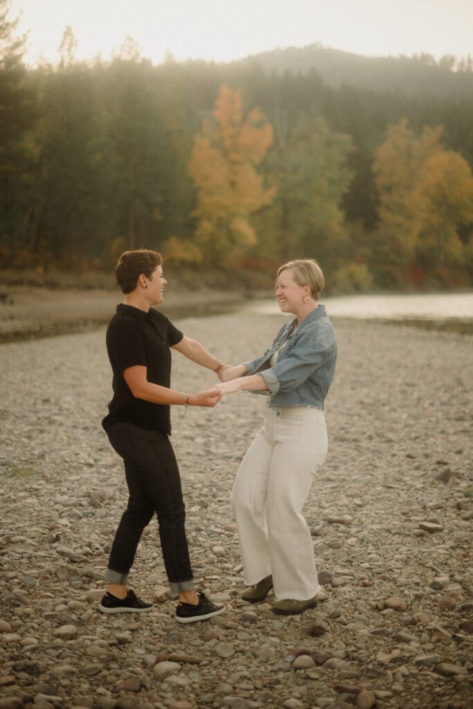 Lesbian Proposal in Missoula: Maclay Flat in Autumn. Photo of two women kissing after getting engaged. On the rocky shore line with trees surrounding them. 