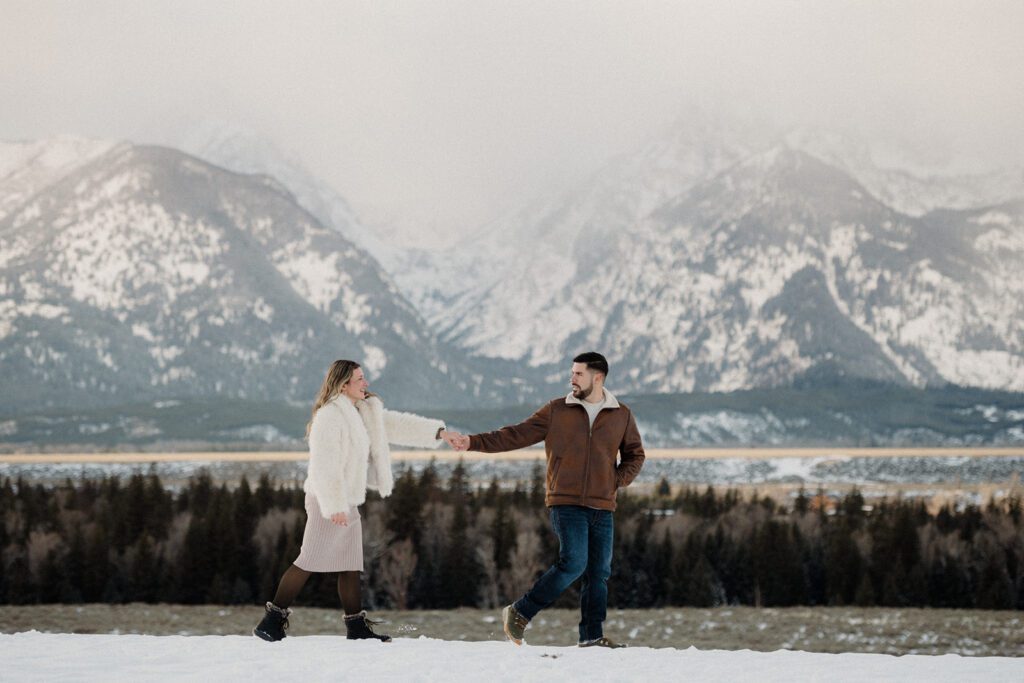 Proposal in Grand Teton with mountains behind newly engaged couple after a snowy proposal. Man holding his lady's hand as they explore. 