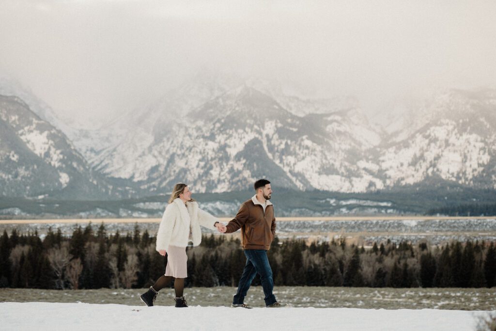 Proposal in Grand Teton with mountains behind newly engaged couple after a snowy proposal. Man holding his lady's hand as they explore. 