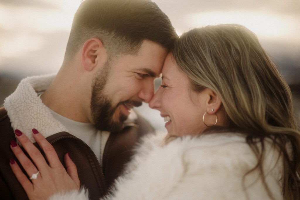 Couple with foreheads pressed together and noses touching. Woman smiling. Engagement ring is prominent on left hand,