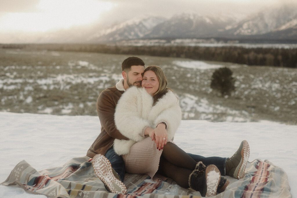 Couple on a blanket together in the snow after getting engaged. He is holding her from behind as she shows off her engagement ring.