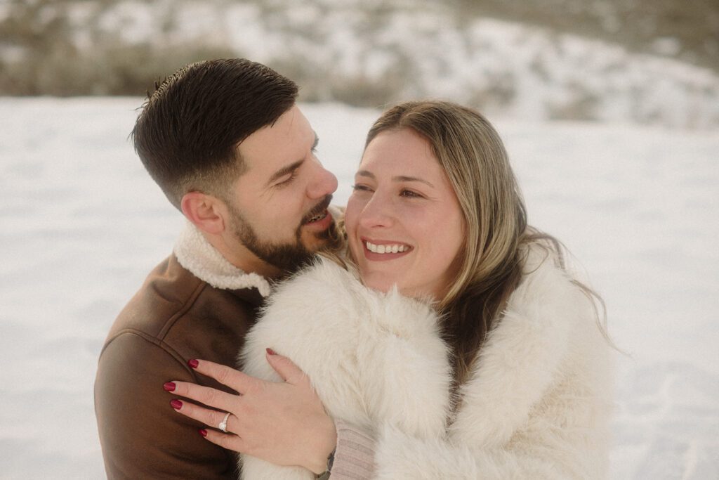 Couple on a blanket together in the snow after getting engaged. He is holding her from behind as she shows off her engagement ring.