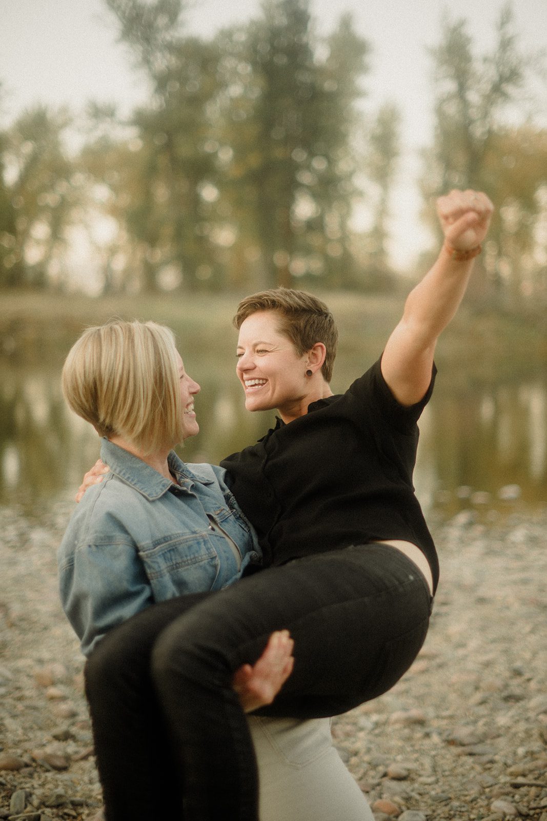 Lesbian Proposal in Missoula: Maclay Flat in Autumn. Photo of two women kissing after getting engaged. On the rocky shore line with trees surrounding them. 