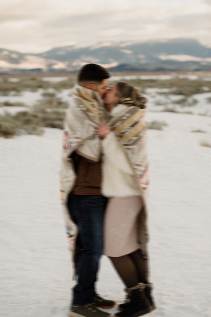 Blurred photo of man and woman kissing in the snow with a blanket around them. 