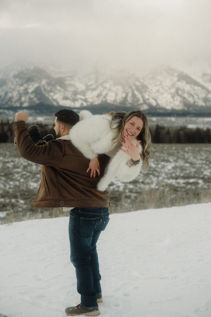 Proposal in Grand Teton. Man in brown  jacket carrying new fiance over his shoulder. He has a fist up in the air in celebration.