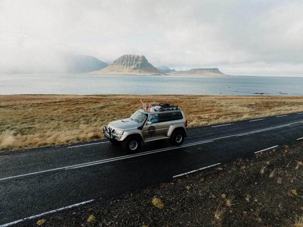 Iceland Queer Elopement, lesbian couple in a jeep with heads out the sun roof, driving along the water.