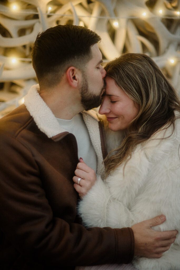 Proposal in Grand Teton. Man kissing new fiancés forehead under twinkling lights in Jackson Hole, WY.