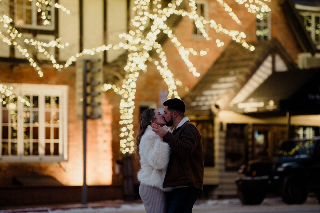 Couple kissing during winter wonderland engagement, holding hands underneath twinkling lit tree. 