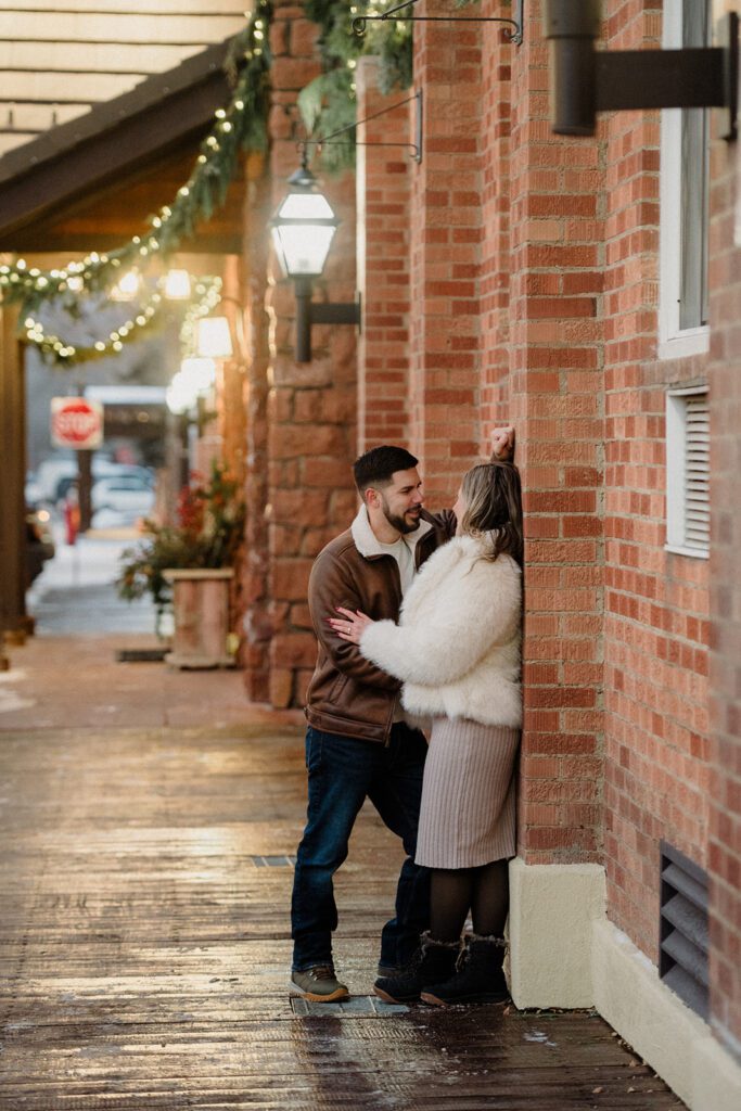 Newly engaged couple kissing against a brick wall, dressed in winter clothing in downtown Jackson Hole, Wyoming. 