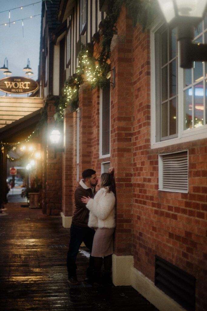 Newly engaged couple kissing against a brick wall, dressed in winter clothing in downtown Jackson Hole, Wyoming. 