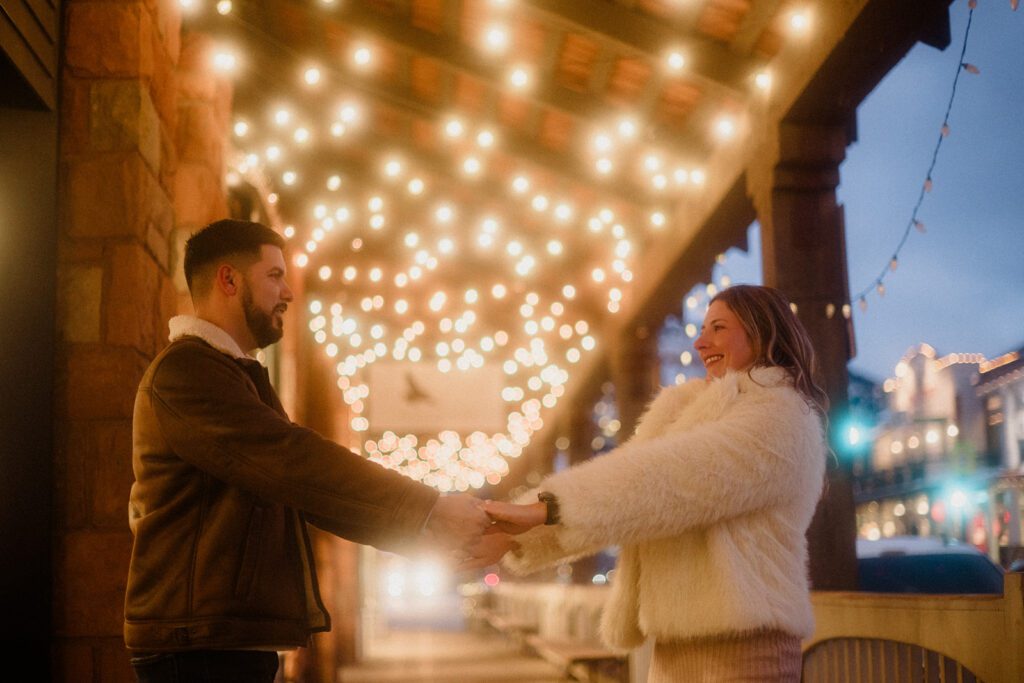 Man and woman in Downtown Jackson Hole holding hands under twinkling lights, celebrating a winter proposal.