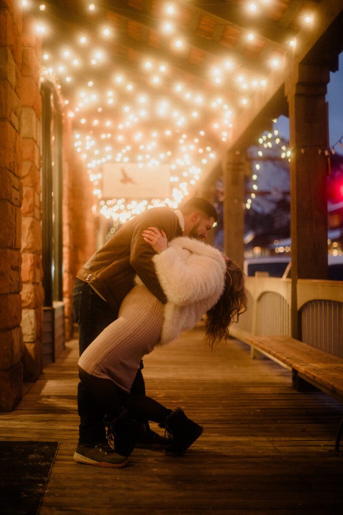 Man dipping woman in Downtown Jackson Hole under twinkling lights, celebrating a winter proposal.