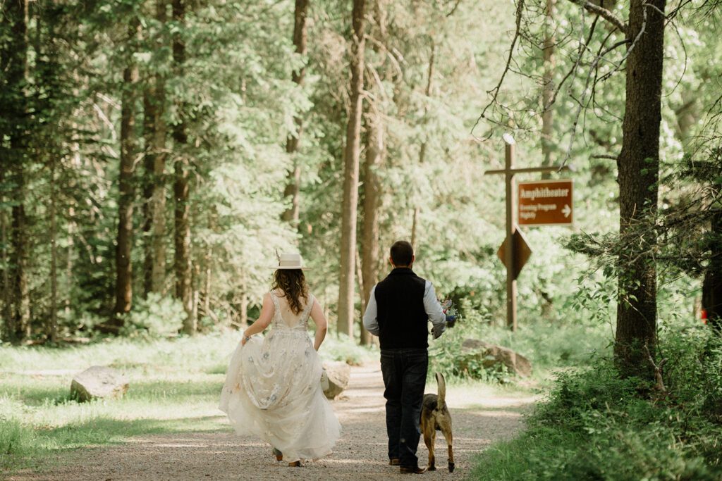 Photo of the back of the Bride and Groom walking toward Apgar Theater in Glacier National Park. Green trees surround them.