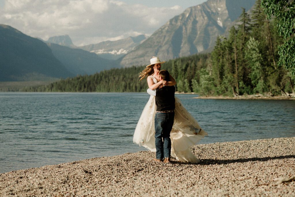 How to have a micro wedding in Glacier National Park with 30 friends and family. Groom picking up and spinning bride on the shores of Lake McDonald.