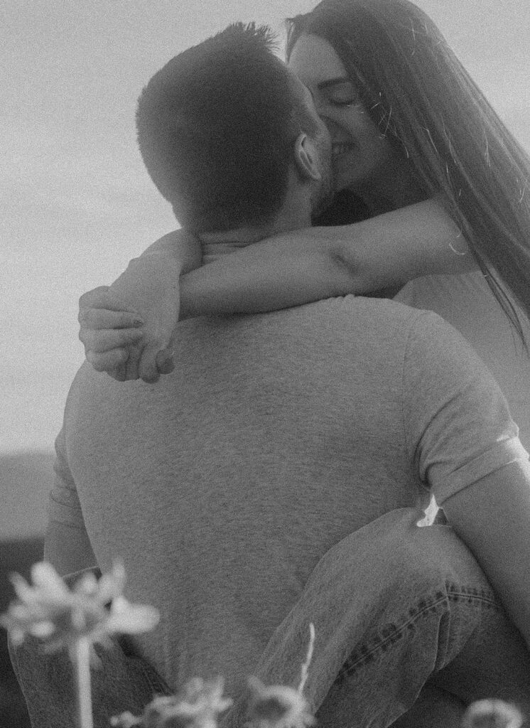 Black and white photo of man holding woman in a flower field while kissing her.