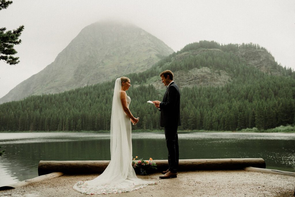 Groom reading vows to wife during their National Park wedding in Glacier! Lake with raindrops behind them.