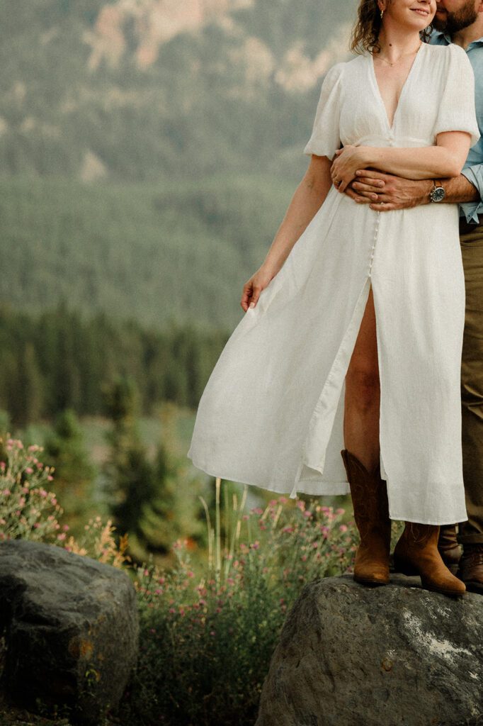 Bozeman dog friendly engagement photos on bridge. Man holding laughing woman from behind. Beautiful white dress with a slit.